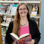 MPHPL Library Associate Janna McLean holds a book at the downtown Mishawaka Library and smiles.