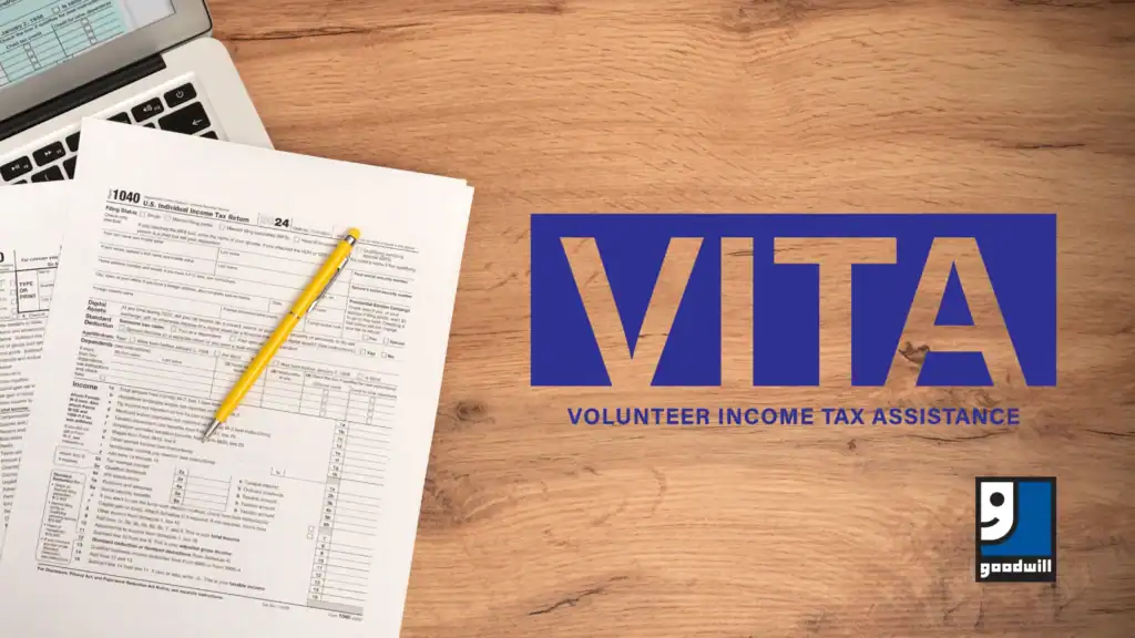Goodwill logo. Tax forms and a yellow pencil on a wooden desk next to a laptop. 'VITA Volunteer Income Tax Assistance.'
