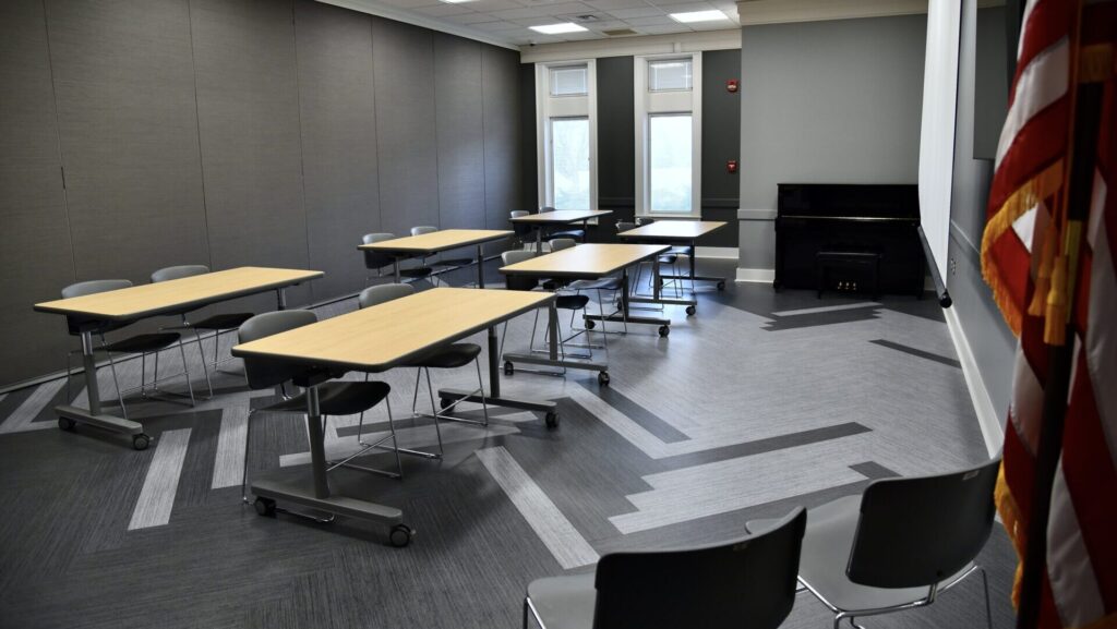 A modern classroom with gray walls, several light wood tables and gray chairs arranged in small groups, large windows letting in natural light, a piano against the wall, and part of an American flag visible in the corner.