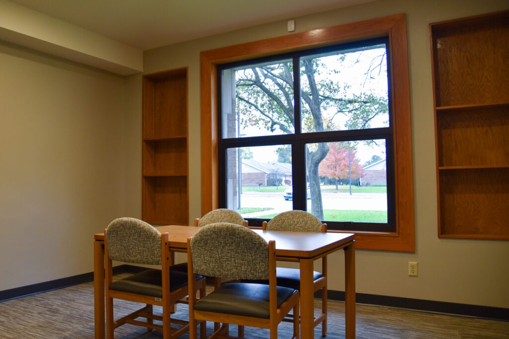 A small study room with a wooden table and four chairs, two empty bookshelves, and a large window showing trees and a street outside. Natural light brightens the neutral-colored space.
