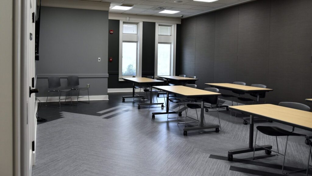A modern, empty classroom with gray walls, several tables, and chairs arranged in rows. Two large windows let in natural light, and extra chairs are stacked in the corner. The room has a clean, minimal design.
