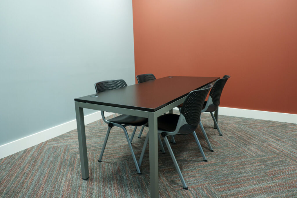 A small conference room with a rectangular black table and four black chairs on a patterned carpet, with light blue and reddish-brown walls.