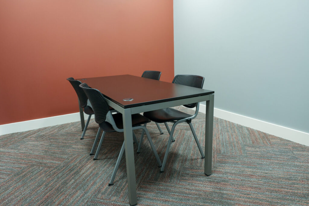 A minimalist meeting room with a black rectangular table and four black chairs, set on a patterned carpet. The walls are painted muted red and light gray, with white baseboards.