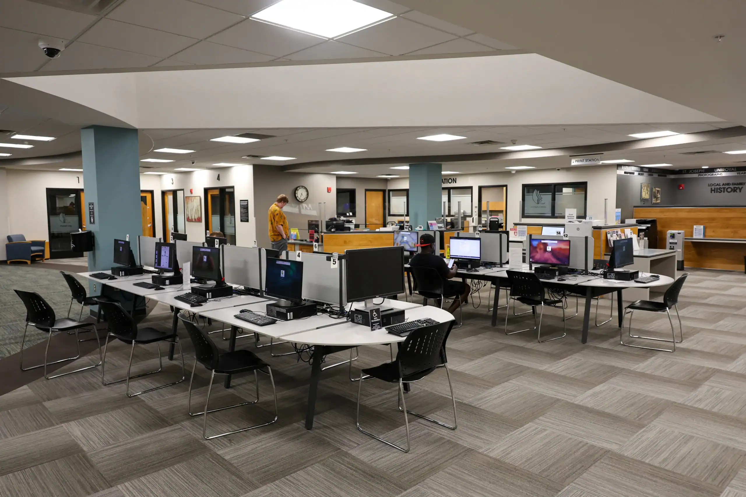 A spacious library computer lab with multiple desks and computer stations, a few people working, and bookshelves and service counters in the background. The room is well-lit with modern decor.