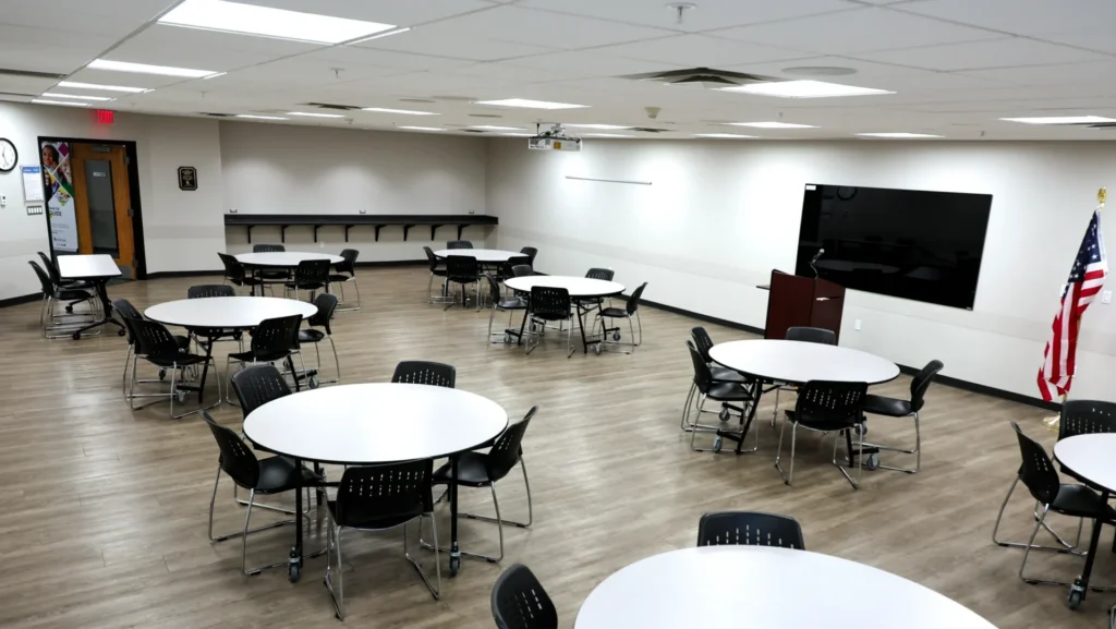 The Mishawaka Library Eisen Meeting Room containing a dry erase board, a podium and rows of circular tables, each surrounded by chairs. The room has light wood flooring, white walls, and a modern appearance.