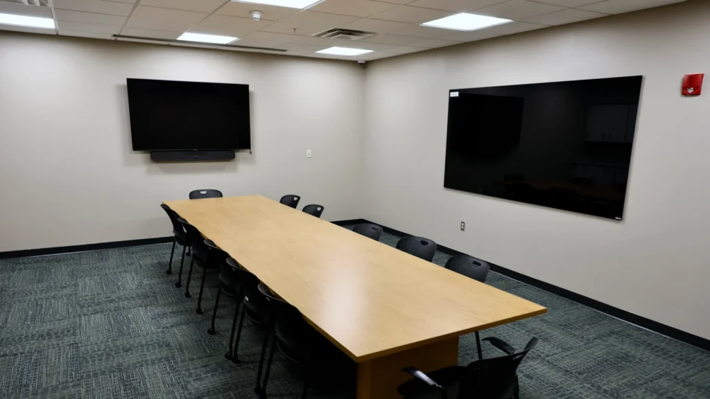 The Mishawaka Library Friends Meeting Room containing a large wall mounted TV and speaker on the left wall, a large dry erase board on the right wall, and a long rectangular wooden table centered in the room, surrounded by chairs. The room has dark carpeted flooring, grey walls, and a modern appearance.