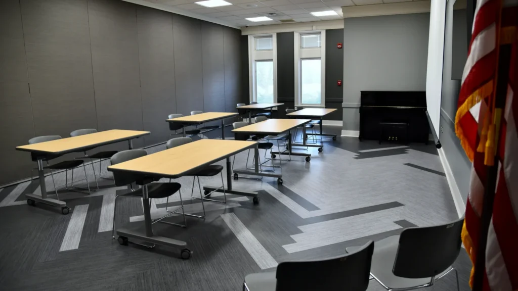 The Harris Branch Meeting Room B containing six tables that seat two chairs, a large wall mounted TV, a room divider wall, and a piano. The room has vinyl flooring, grey walls, and a modern appearance.