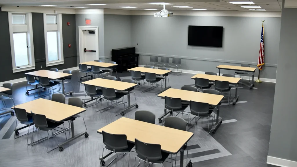 The Harris Branch Meeting Room A and B, without a divider wall, containing nine tables that seat four chairs, a large wall mounted TV, and a piano. The room has vinyl flooring, grey walls, and a modern appearance.