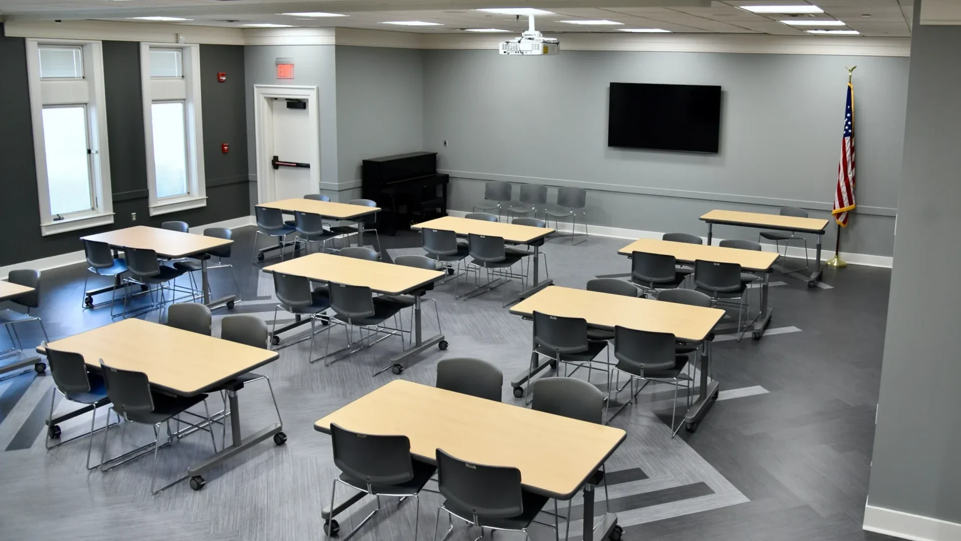 The Harris Branch Meeting Room A and B, without a divider wall, containing nine tables that seat four chairs, a large wall mounted TV, and a piano. The room has vinyl flooring, grey walls, and a modern appearance.