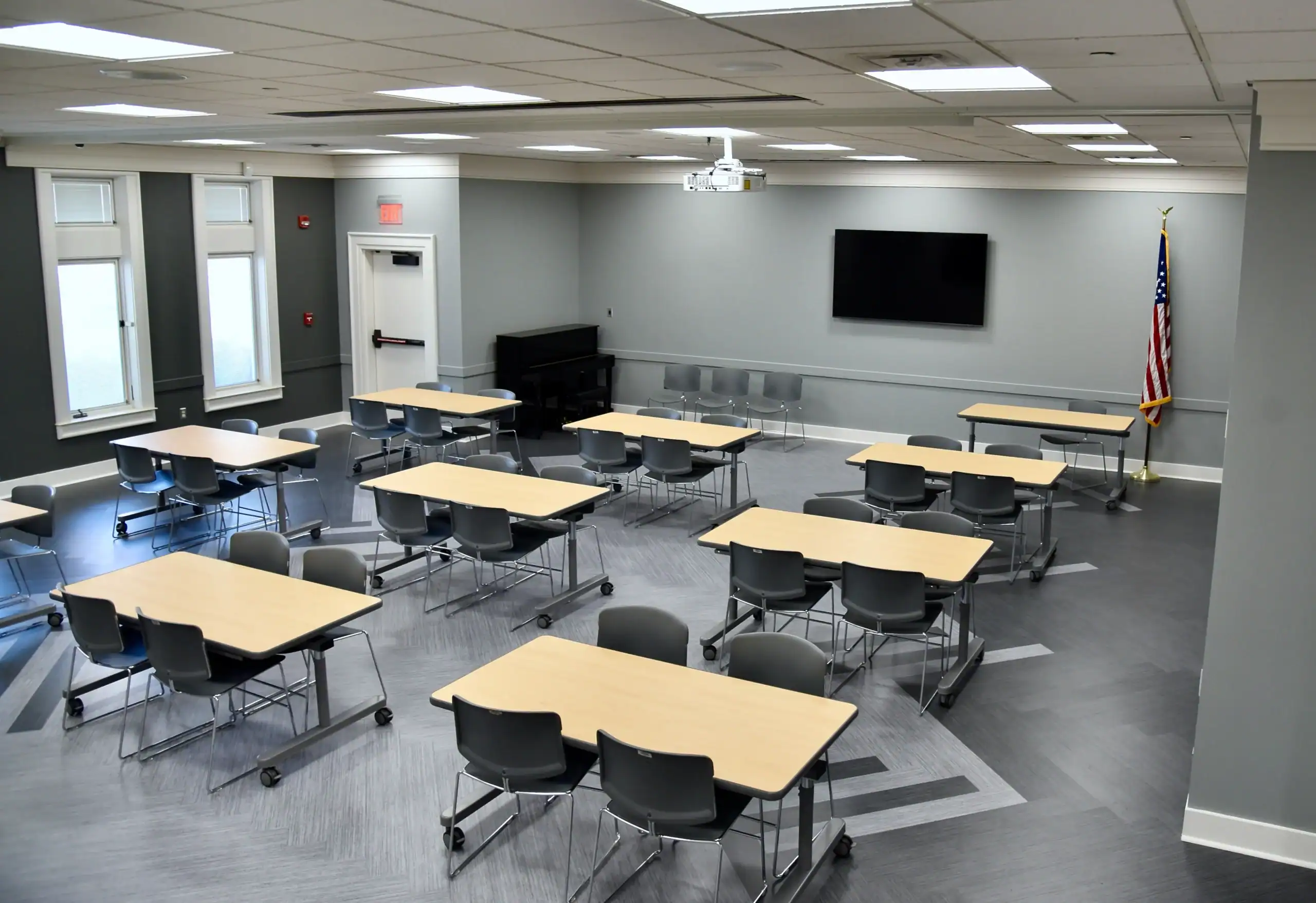 A modern classroom with several rectangular tables and gray chairs, arranged in small groups. There is a large wall-mounted TV, windows, an American flag, and a piano in the corner. The room is empty and well-lit.