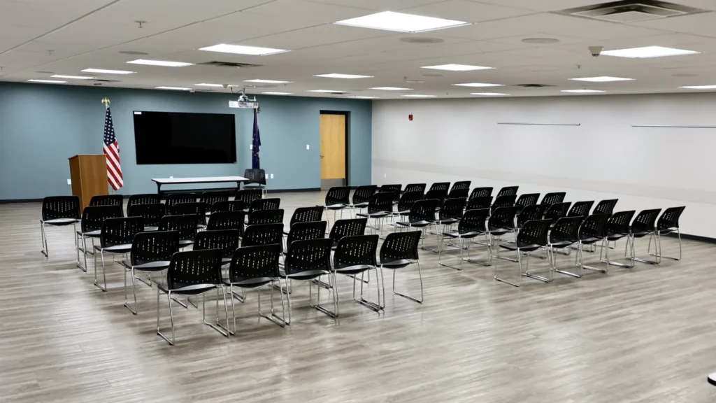 The Mishawaka Library Spencer Meeting Room containing rows of black chairs facing a podium, two flags, and a large dry erase board. The room has light wood flooring, white and blue walls, and a modern appearance.