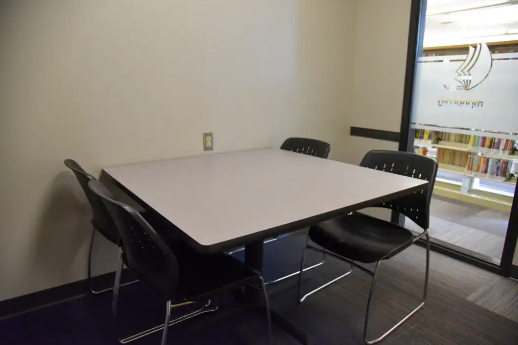 The Mishawaka Library Youth Study Room One containing a table with four chairs. The room has a plain wall and a window showing bookshelves outside.