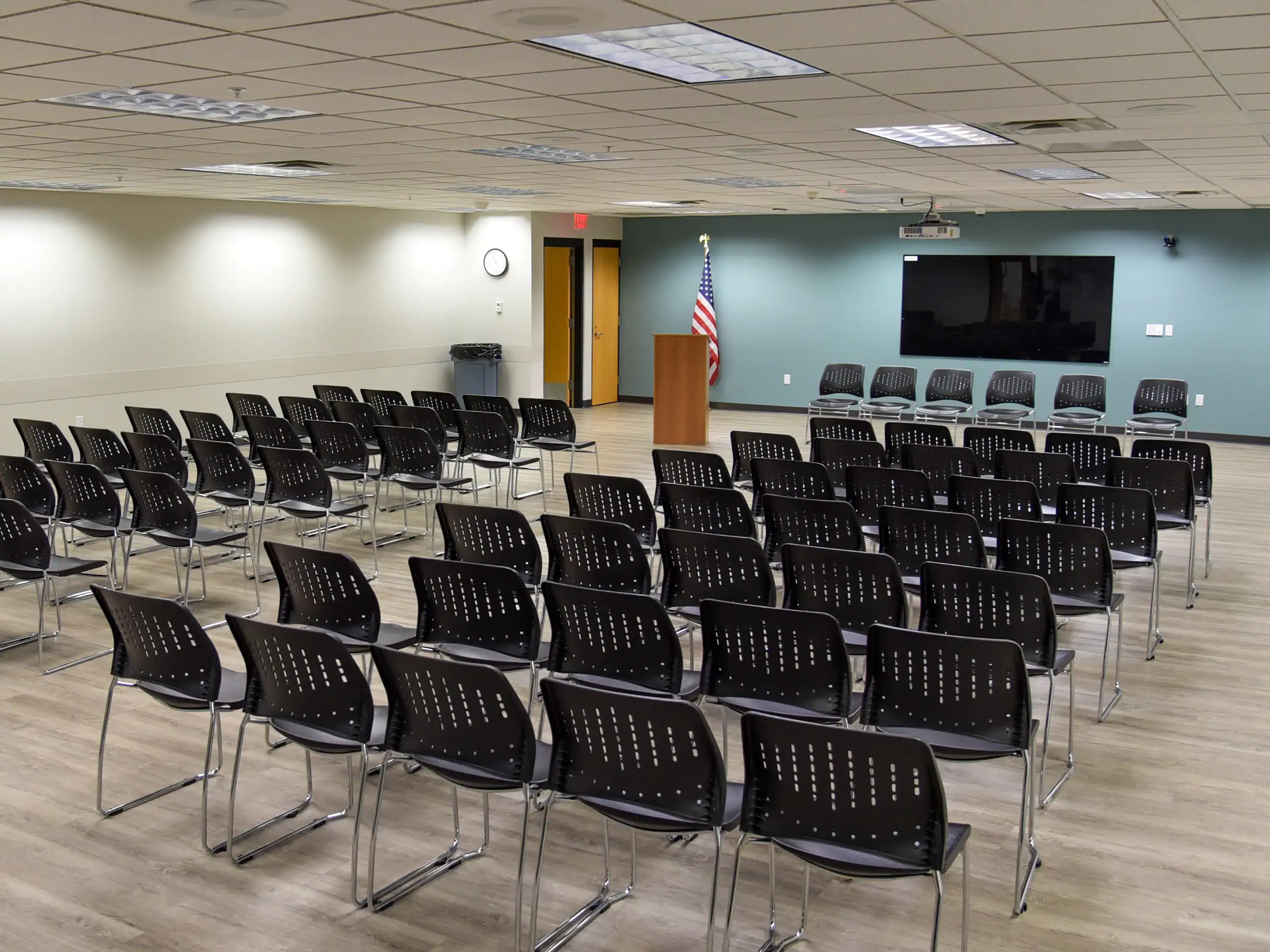 Rows of empty black chairs face a podium with an American flag in a modern, well-lit conference room. A large TV screen is mounted on the wall behind the podium, and the floor is wood.