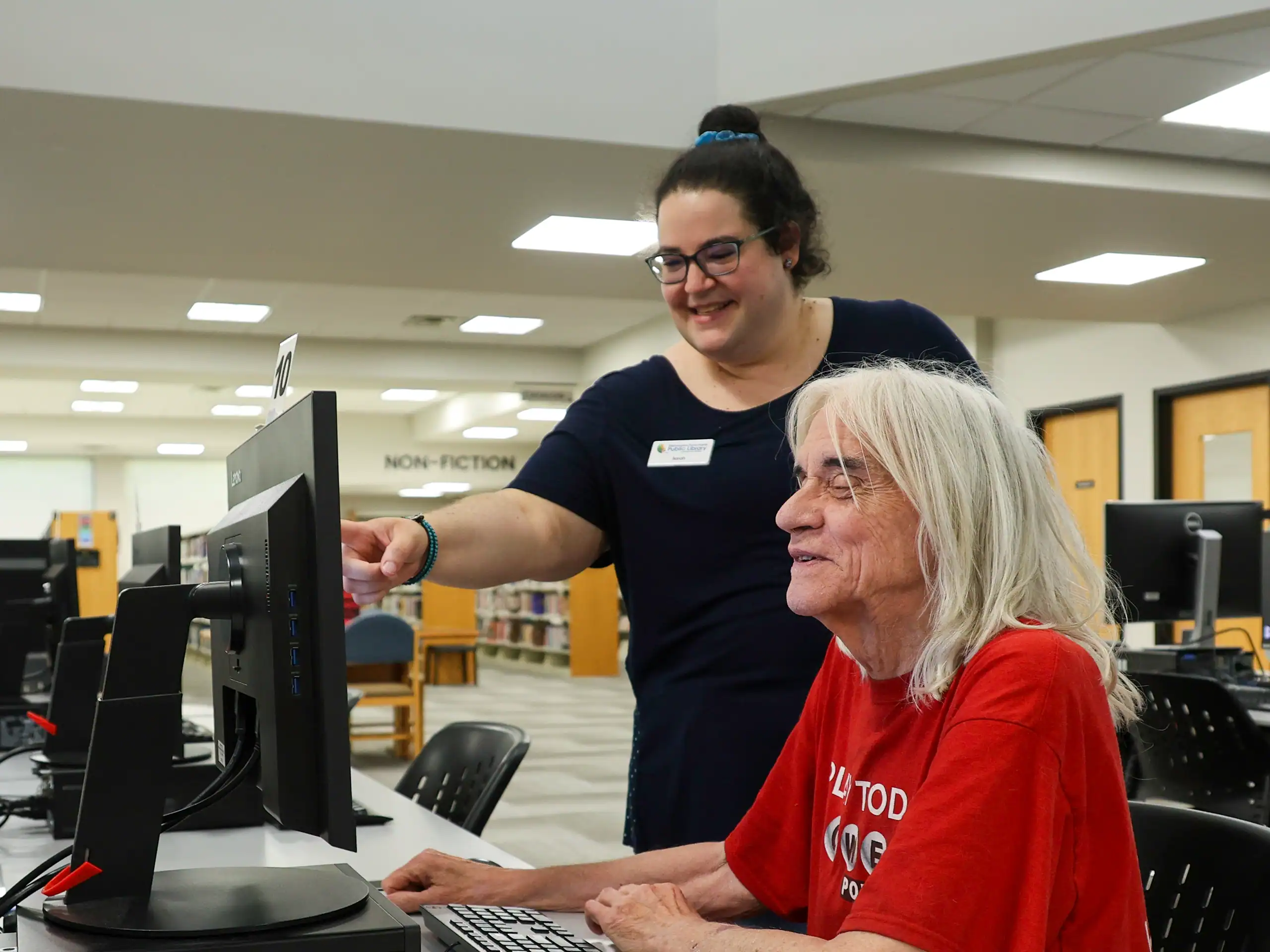 A woman in a red shirt sits at a computer at the Mishawaka Library, smiling, as a staff member standing beside her points at the monitor, offering assistance. Other computers and desks are visible in the background.