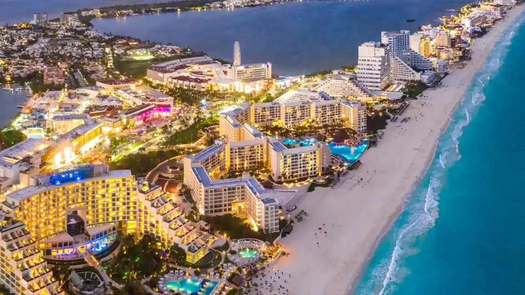 Aerial view of a vibrant beachfront resort featuring pools, hotels, and city lights by the ocean.