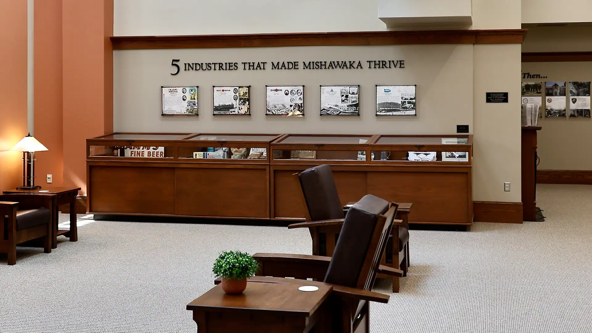A museum exhibit titled 5 Industries That Made Mishawaka Thrive, with display cases, framed industry posters on the wall, and wooden chairs and tables in a carpeted, well-lit room.