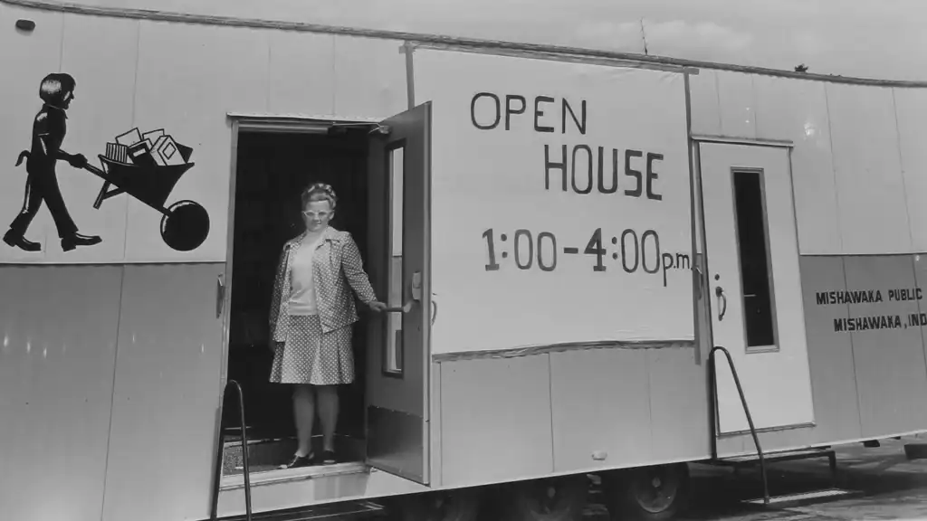 A woman stands in the doorway of a mobile building with a large “Open House 1:00-4:00 p.m.” sign. The building has a painted child with a wheelbarrow and a sign for Mishawaka Public Schools, Mishawaka, Indiana.