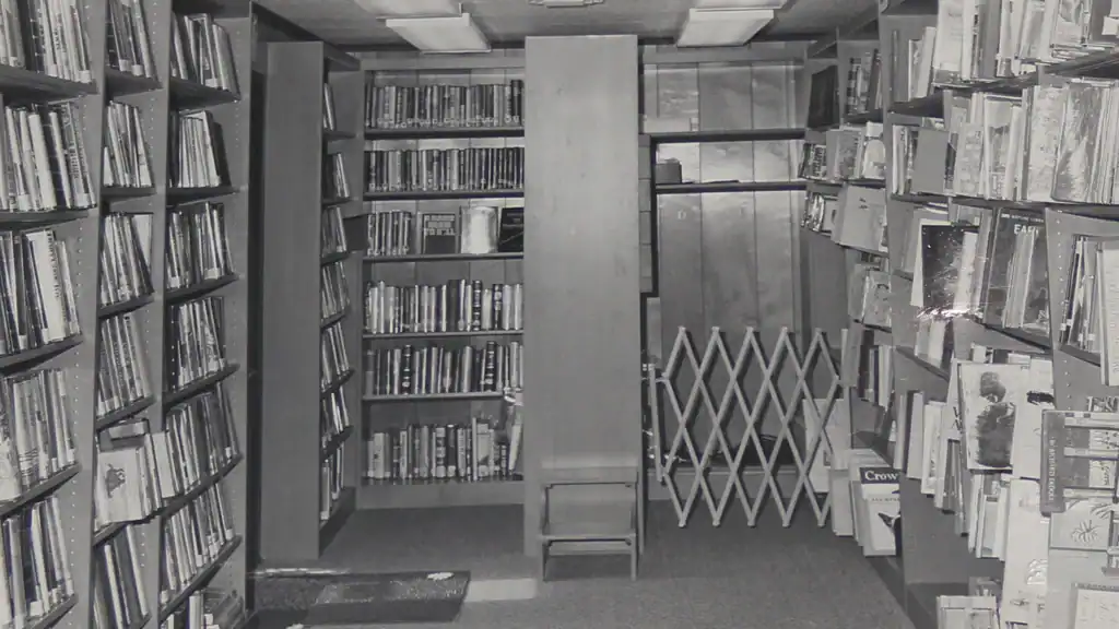 Black and white photo of a narrow library aisle with tall bookshelves on both sides, a small step stool in the center, and a metal accordion gate at the back near a closed wooden door.