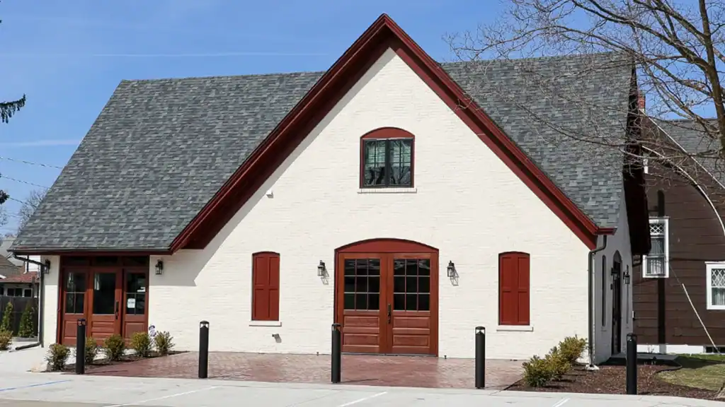 A cream-colored building with dark red trim, steep gray shingle roof, and double wooden doors, situated by a brick walkway, with a few small bushes and leafless tree branches nearby.
