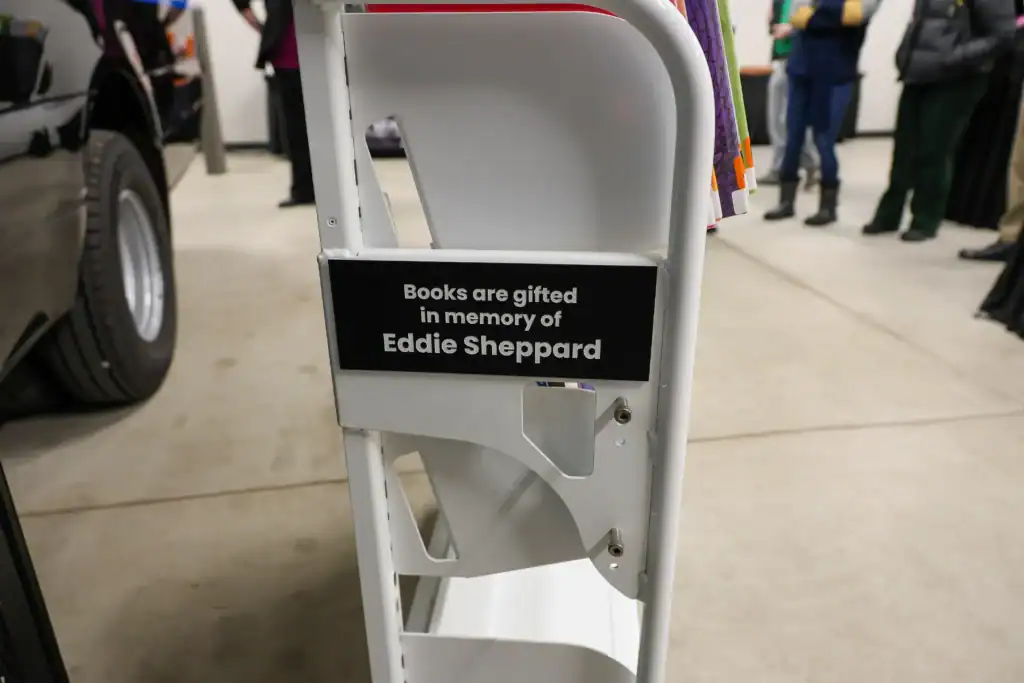 White book cart with a sign reading ‘Books are gifted in memory of Eddie Sheppard.’ People in the background at the MPHPL Bookmobile launch.