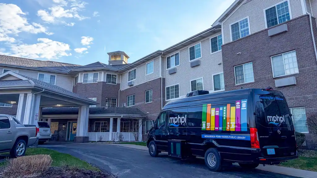 The MPHPL Bookmobile with colorful book graphics is parked outside a multi-story brick and siding building under a partly cloudy sky. Nearby, other vehicles are parked by the entrance.