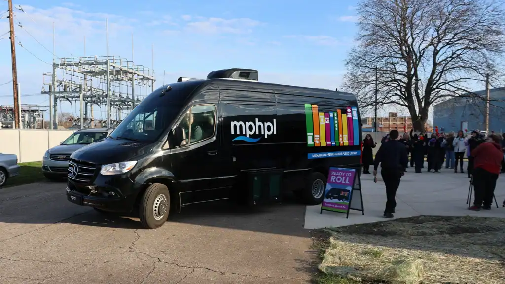 A black van with mphpl and colorful books on the side is near a sign that reads 'ready to roll'. People cheer and take photos, conveying excitement. A cameraman films the scene.