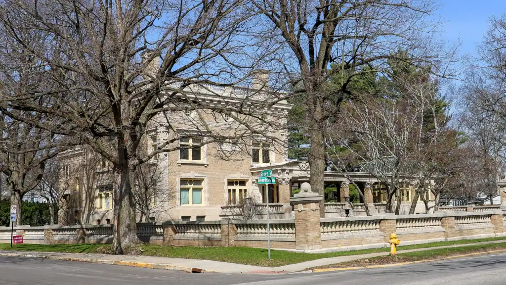 A large historic brick mansion with tall windows and a wraparound porch sits behind leafless trees and a stone fence on a street corner with a yellow fire hydrant and street signs visible.