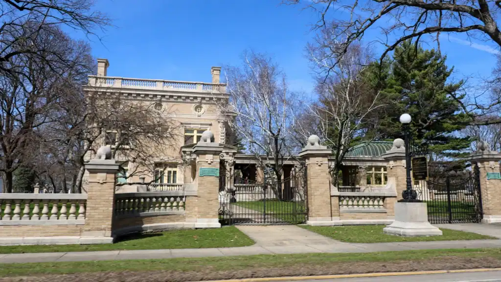 A large, historic mansion with ornate details and a balcony sits behind a stone and iron fence, surrounded by leafless trees and green grass under a clear blue sky.
