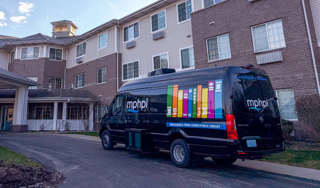 The MPHPL Bookmobile with colorful book graphics and logo parked outside a large brick multiple story building. The sky is clear and there is some grass beside the drive.