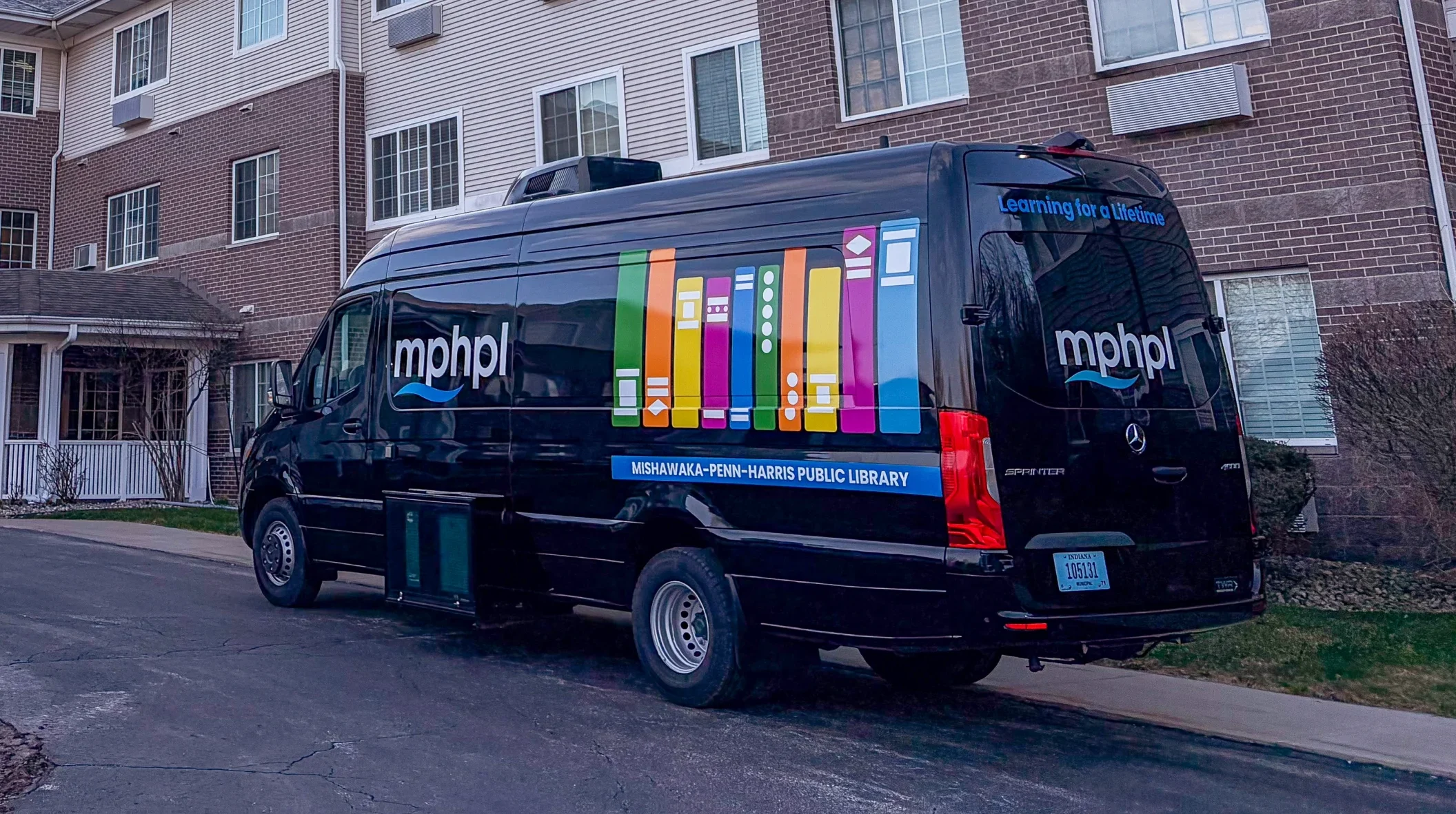 A black Mishawaka-Penn-Harris Public Library van with colorful book graphics is parked outside a multi-story brick and white building, likely making a community stop.