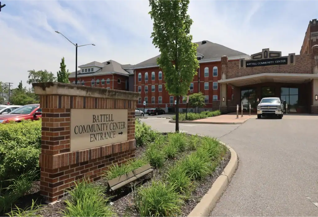 A brick sign reading Battell Community Center Entrance with an arrow stands by a landscaped path; behind it is a large red-brick building labeled Battell Community Center and a parked white van.