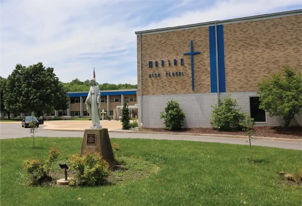 A brick building labeled Marian High School with a blue cross on the wall. A statue stands on a pedestal in front, surrounded by grass, trees, and a sidewalk under a partly cloudy sky.