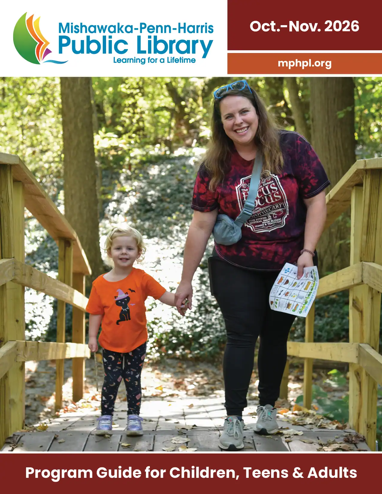 October to November 2026 Program guide cover. ‘MPHPL logo, mphpl.org Program Guide for Children, Teens and Adults'. An adult and child hold hands and smile while walking on a bridge at the Mishawaka Res. They are both wearing Halloween themed shirts.